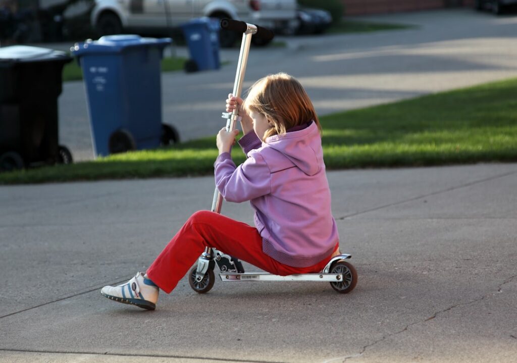 Child riding a scooter on a driveway near garbage bins, emphasizing the importance of garage safety for children.